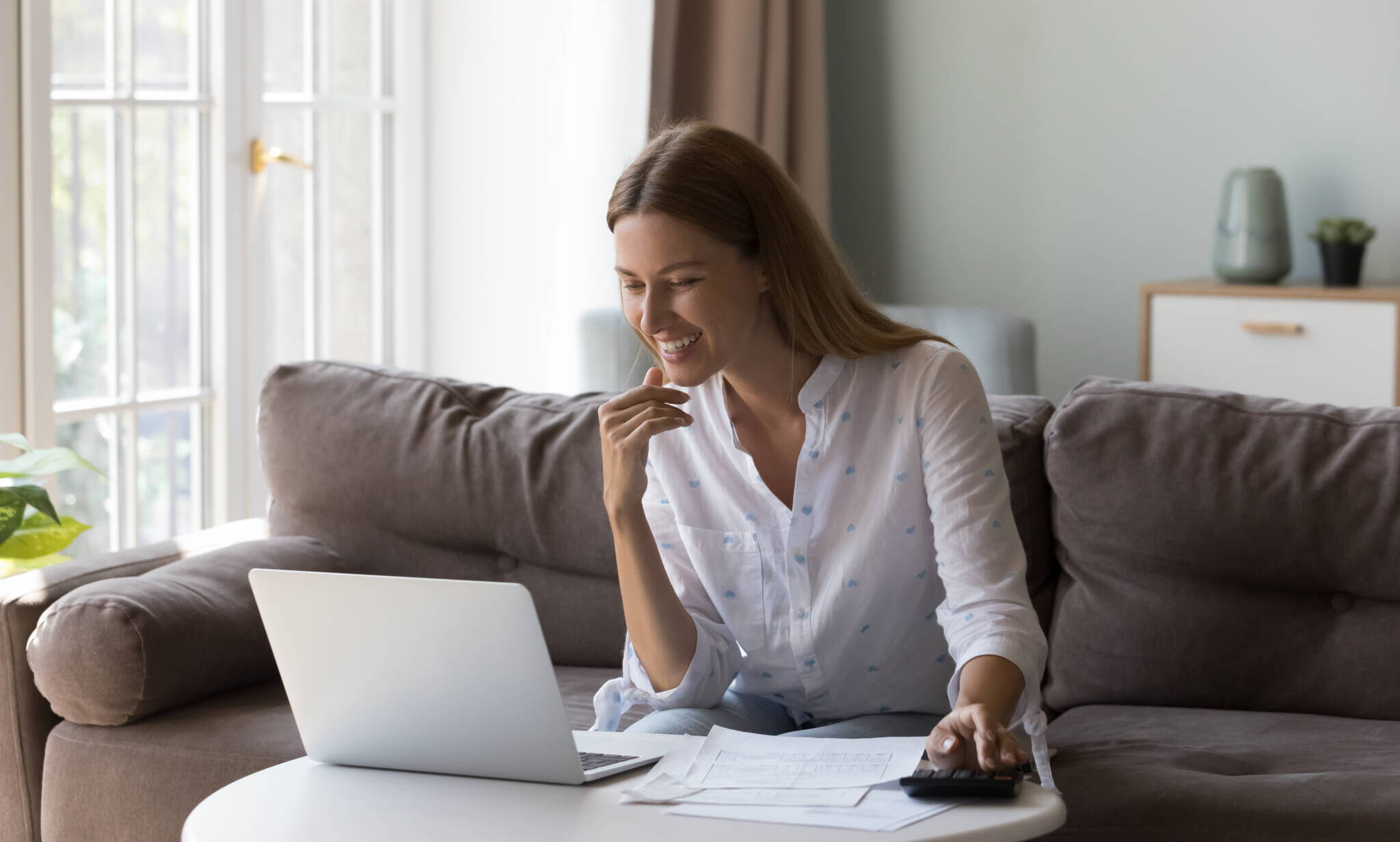Woman Sits At Table With Laptop Counts Incomes Looks Satisfied