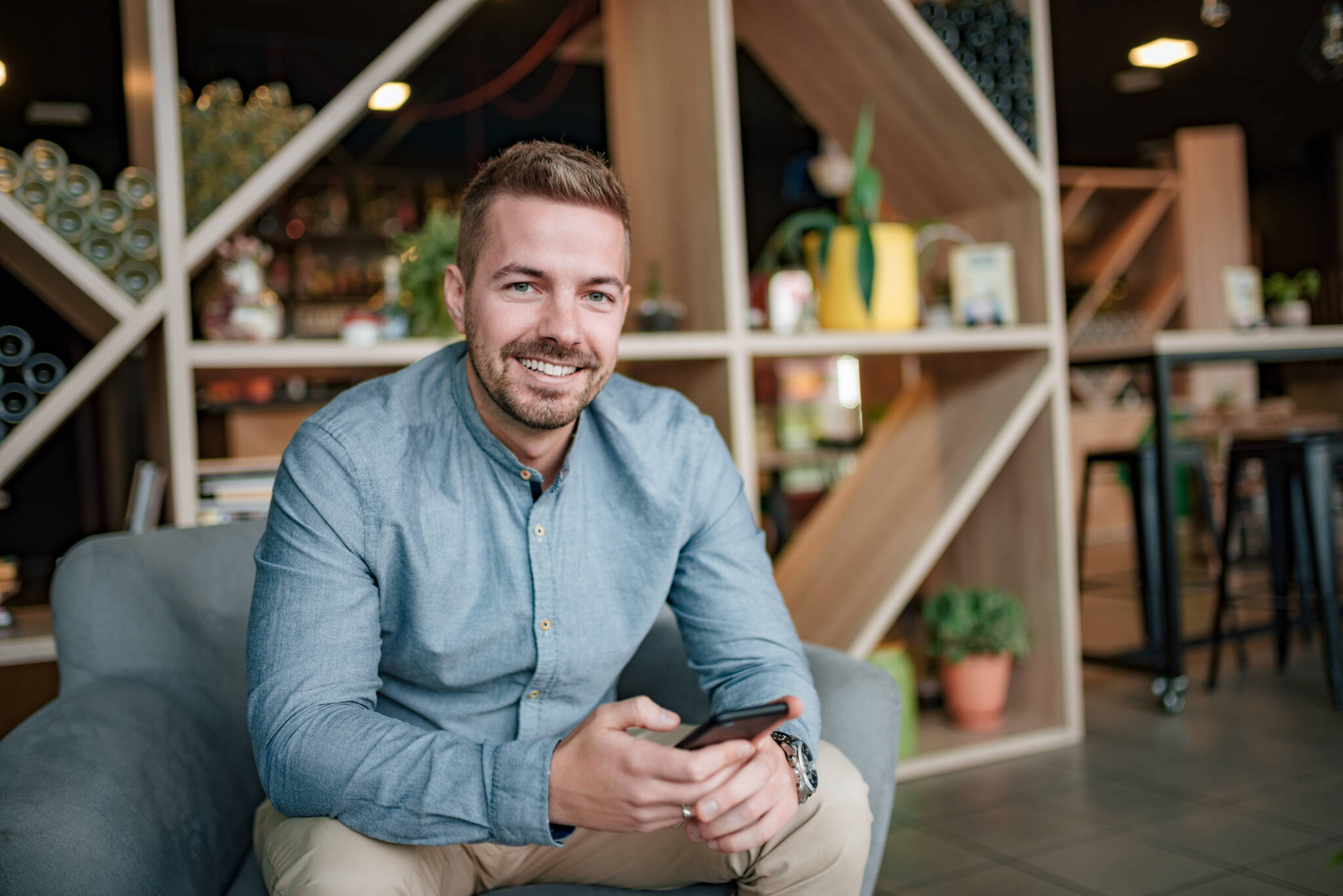 Portrait Of A Handsome Young Man Sitting In A Modern Living Room. Looking At Camera.